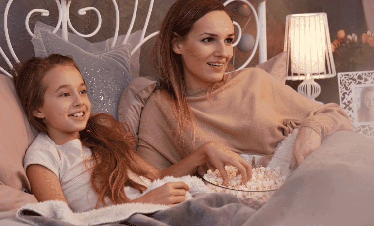 Mother and daughter laying in bed watching a movie together whilst eating popcorn 
