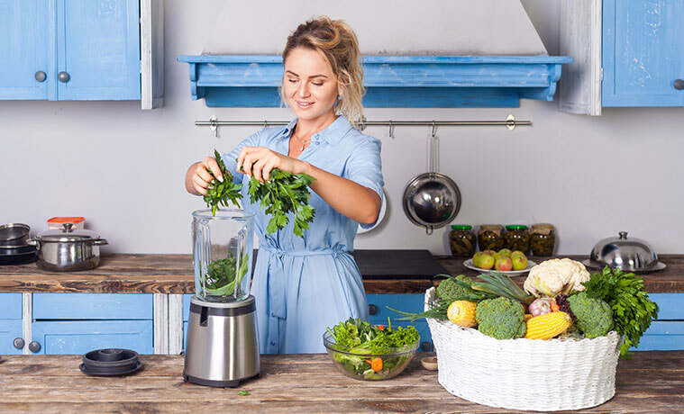 Woman in blue kitchen puts fresh herbs and leafy greens into a silver blender next to basket of fresh vegetables.