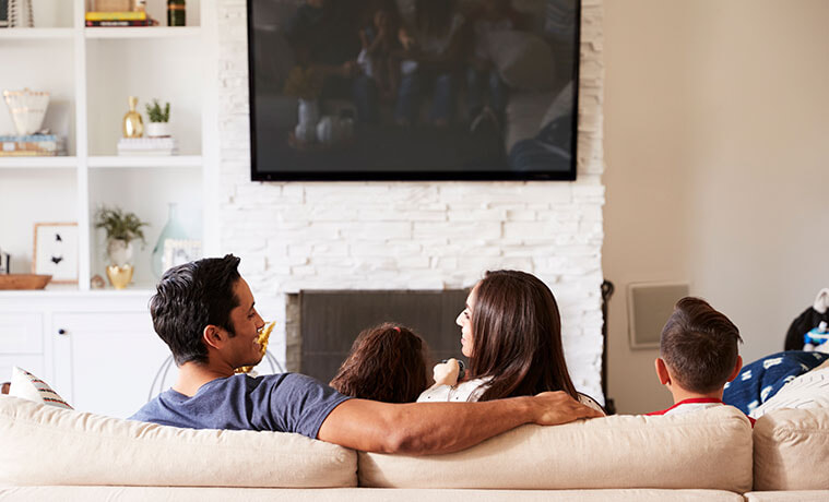 Back view of a young family of four sitting on sofa watching a mounted TV, mum looking at dad.