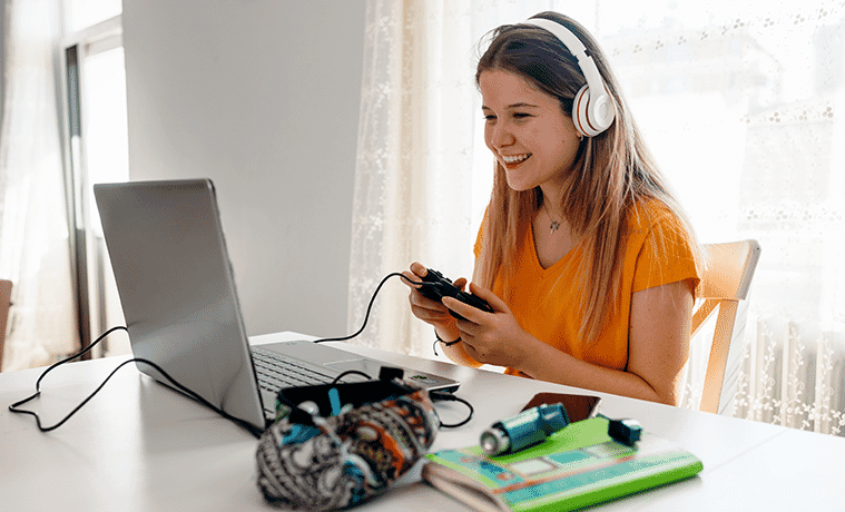 Female uni student with headphones and remote control enjoys a gaming break on a laptop.
