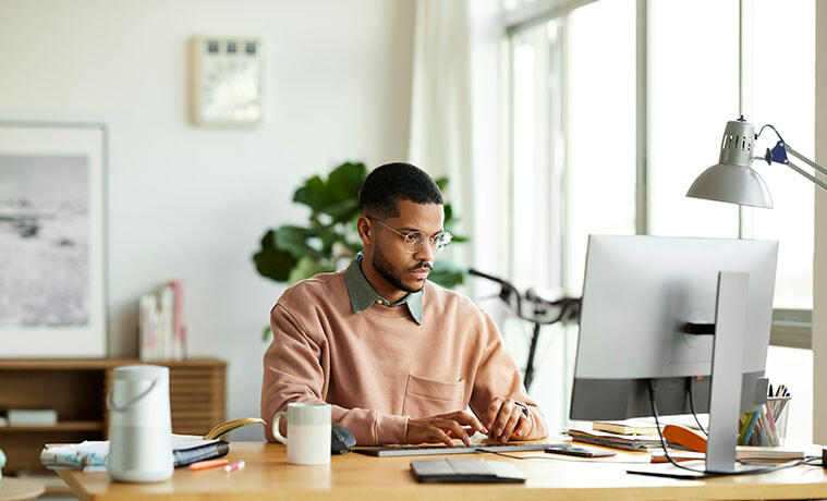 A man sits at the desk in his home office to type notes into his personal computer.