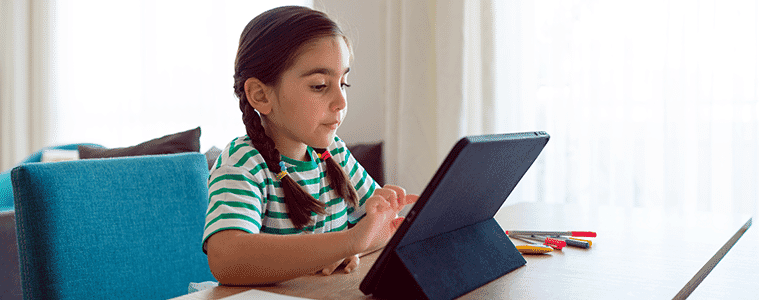 A young girl presses the touchscreen of her tablet while doing homework at the kitchen table