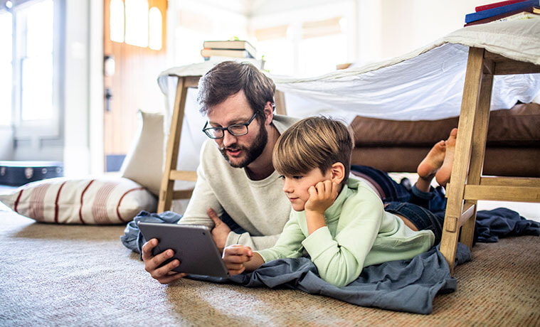 A father and son watch a TV show on their digital tablet while sitting under a homemade fort in their living room.