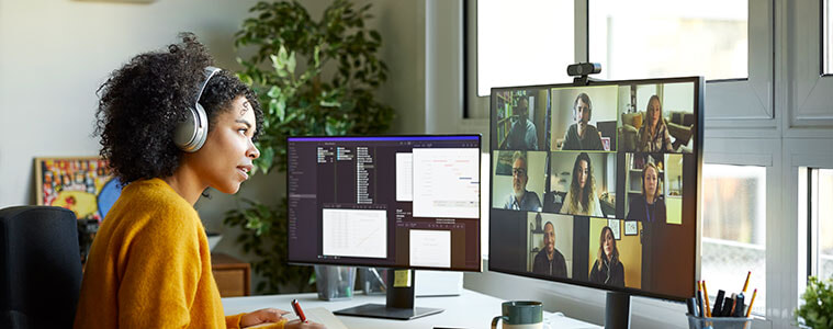 A female entrepreneur, sitting at a desk with two monitors and a keyboard, leads a business meeting on a video call.