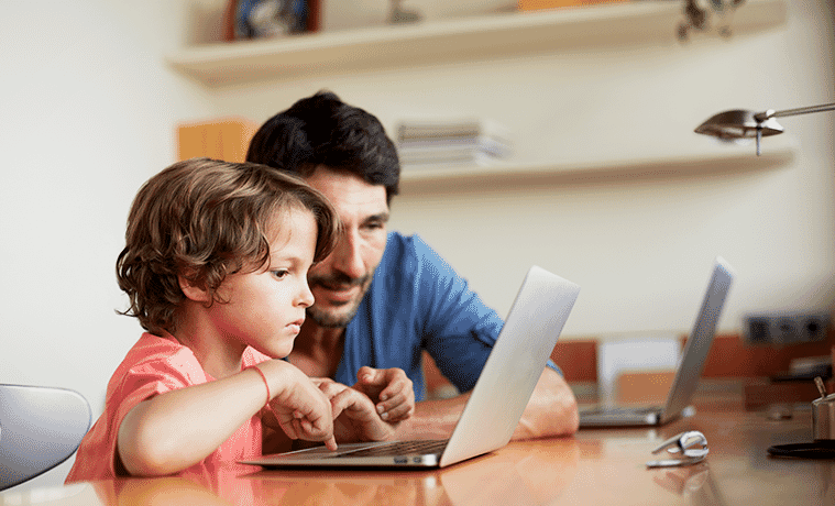 A father and his young son sit together in front of a laptop on a desk in their home office.