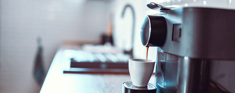 Woman holding a mug of fresh coffee looks at her coffee machine on the kitchen bench. 