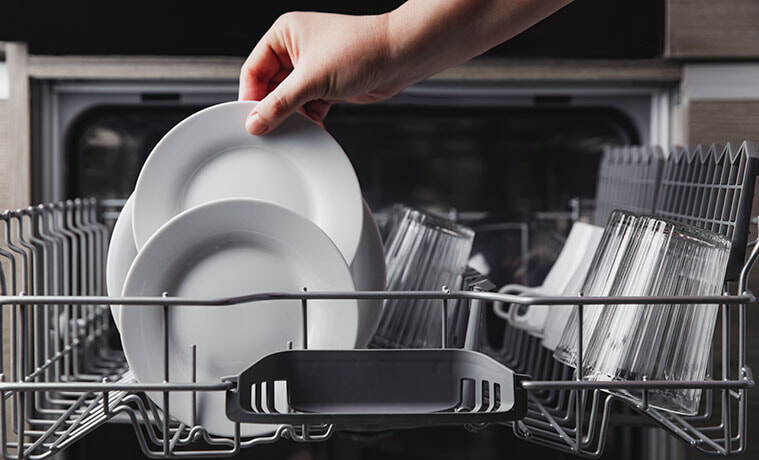 Close-up of a hand removing clean plates and glasses from the top rack in an open dishwasher.