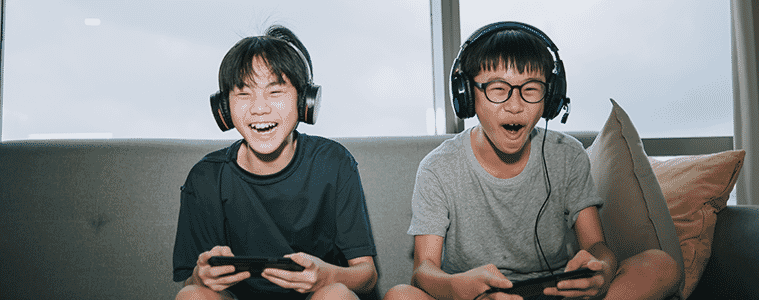 Two brothers on a couch smile while playing video games, each of them holding a gaming console.
