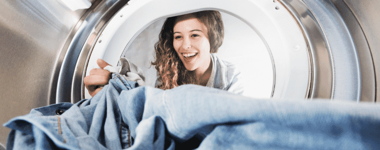 Seen from inside a dryer, a woman reaches into the cavity to remove a pair of jeans. 