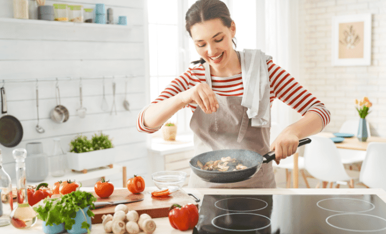 A smiling woman seasons mushrooms in a frying pan she is holding above a sleek cooktop, set into the kitchen bench.