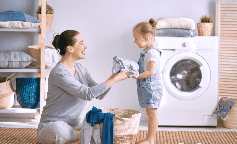 A mother hands her young daughter some freshly dried clothes from a basket in their laundry room.