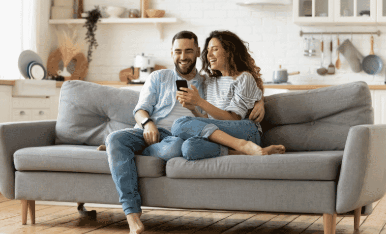 Woman and man snuggle on a couch, laughing while looking at something on the woman’s smartphone screen