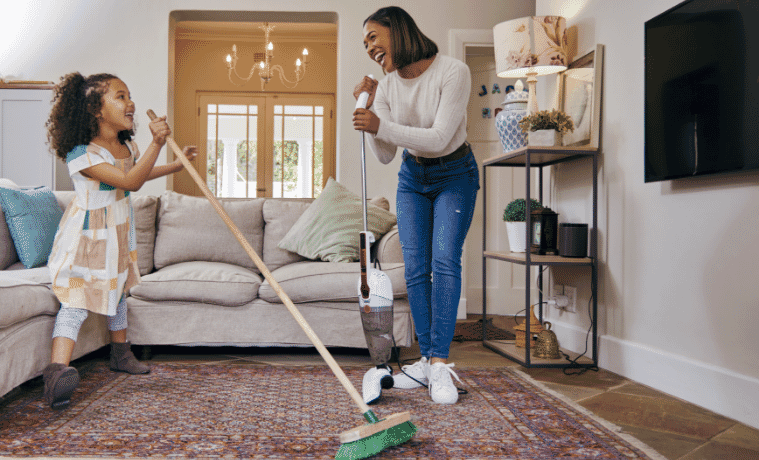 A mother and daughter have fun cleaning the living room with a stick vac and a broom.