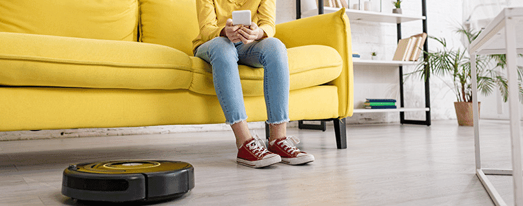 Young girl with a smartphone on a sofa, smiling and looking at a robotic vacuum cleaner on the living room floor.