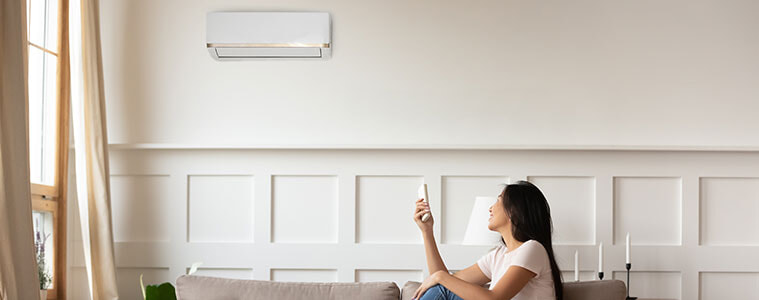 A woman sitting on the couch turns on her wall-mounted air conditioner using a remote control.