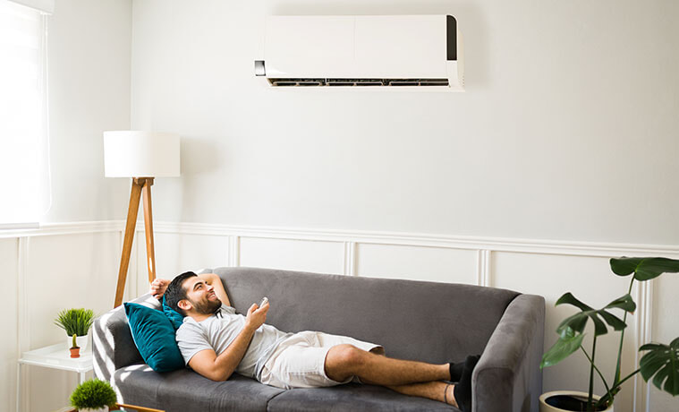 A man lies on the couch in his living room and turns on the wall-mounted air conditioner using a remote control.