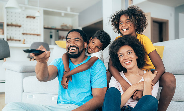 A smiling father holds a black remote and enjoys family viewing time with his wife and children.