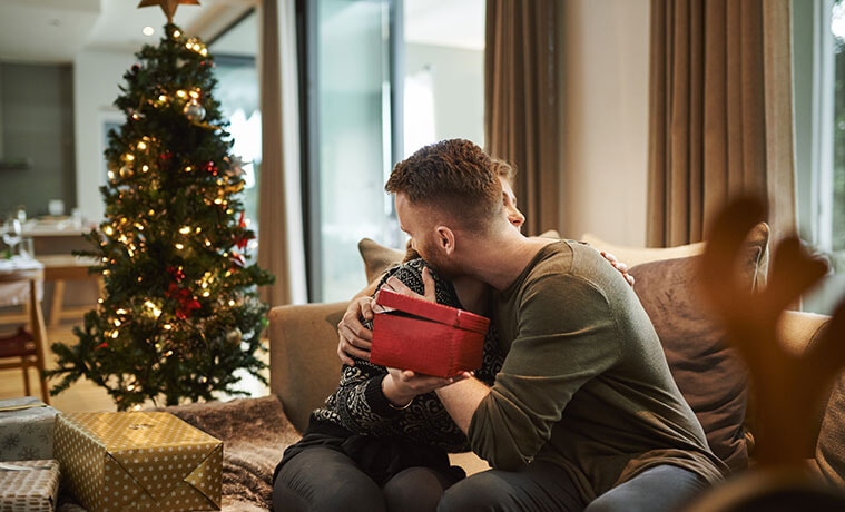 A young couple sitting on a couch near a Christmas tree embrace as they exchange gifts.