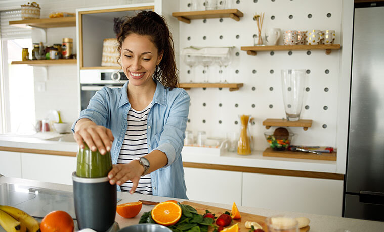 A woman blitzes a green smoothie while standing at her kitchen bench. 