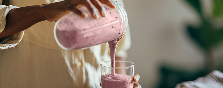 A close-up of a woman’s hands pouring a berry smoothies from a blender cup into a glass at her kitchen bench.