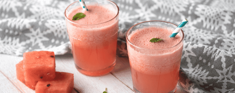 Two glasses filled with watermelon juice with fun straws, and chopped pieces of watermelon and mint sprigs at the side
