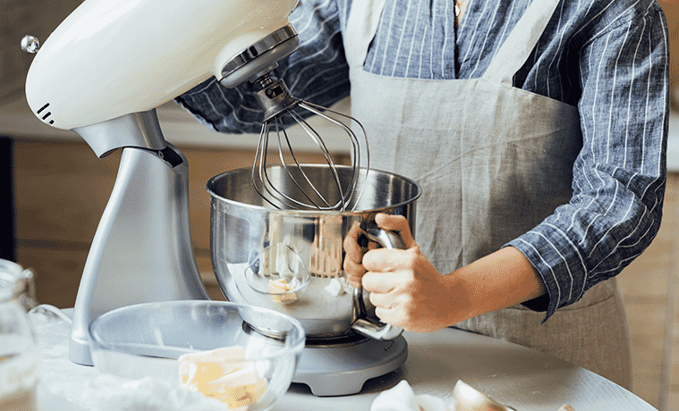 A woman wearing an apron lifts the whisk on her kitchen stand mixer and holds its bowl in her other hand. 