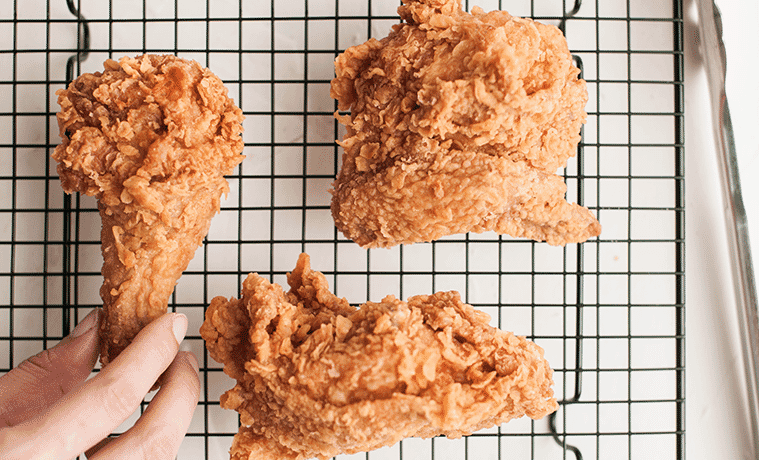 Close-up of a hand reaching for a fried chicken drumstick on a wire rack with two other pieces of fried chicken.