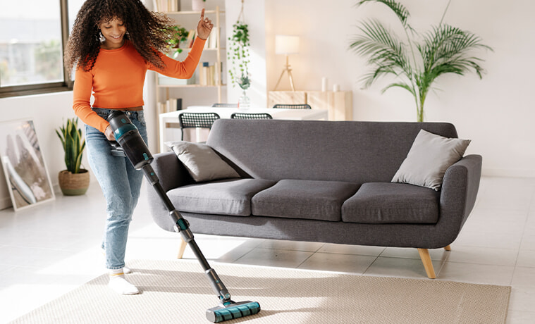 A smiling young woman dances while using the vacuum cleaner in her living room at home