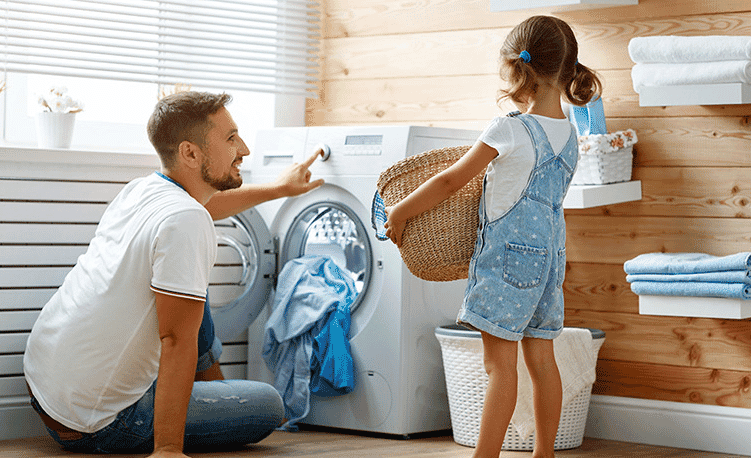 A father and daughter place a load of washing into a front loading washing machine in their laundry.