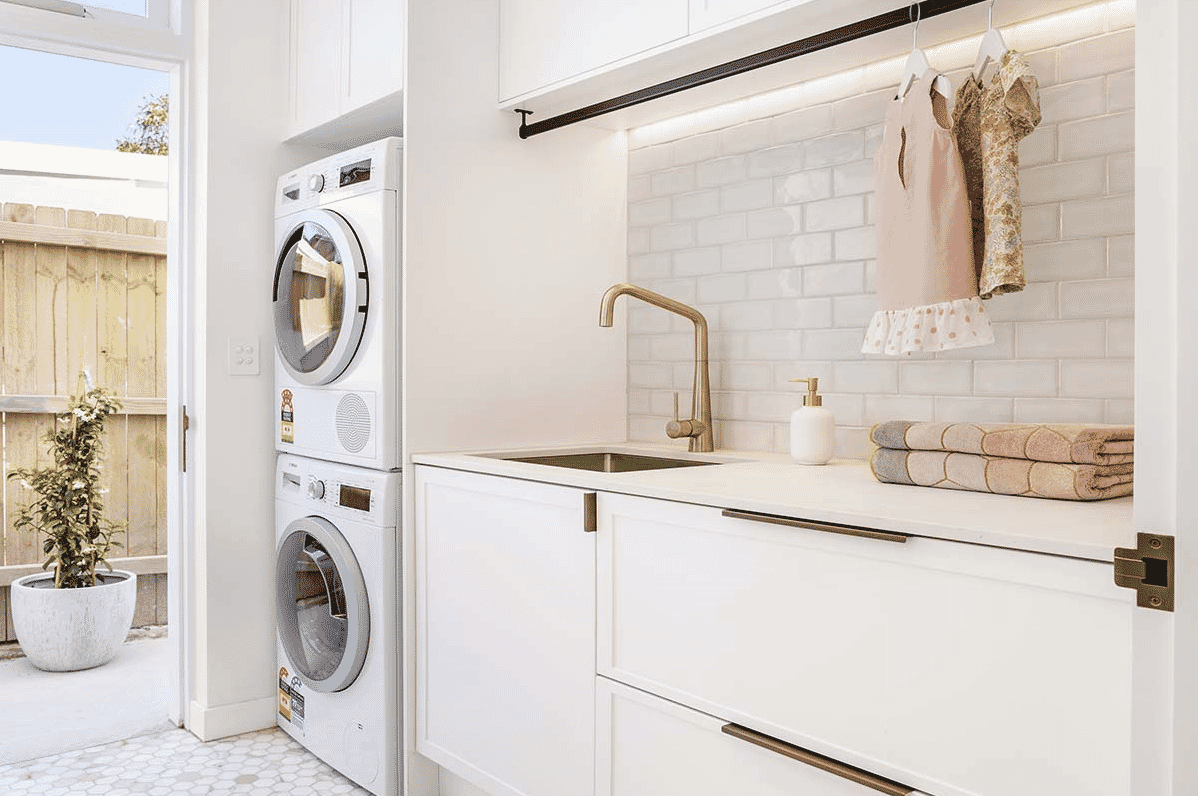Bright sunny laundry with white cabinetry, white subway tiled splashback, brass tapware and stacked washing machine and dryer.