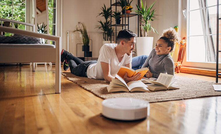 A man and a woman lie on the floor, reading books and chatting, while their robot vacuum cleans the floor.