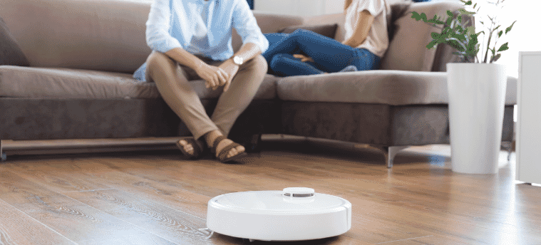 A robot vacuum cleaning the floor whilst a couple sit on a couch in the background