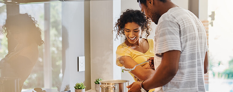 A young couple cooks together in their kitchen, watching a steaming pot sitting on a gas cooktop. 