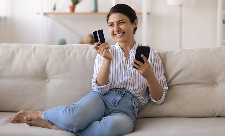Smiling woman sits on the couch at home with her credit card in one hand and her smartphone in the other.