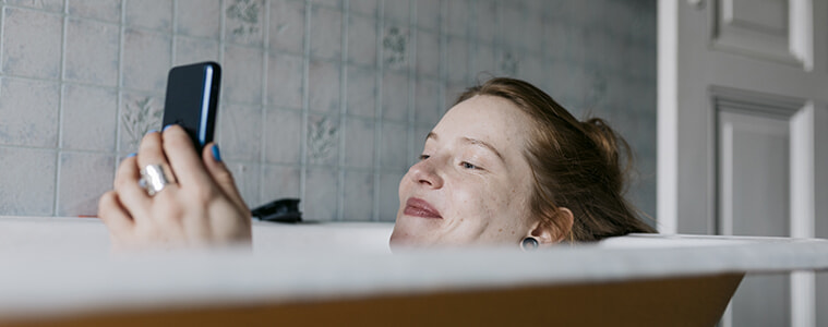 A woman relaxes in the bath while making some online purchases on her smartphone.