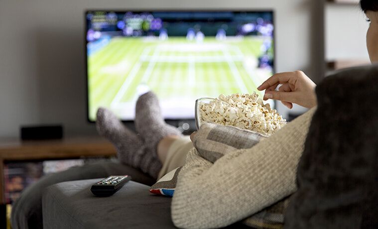 Lady sitting on the couch eating popcorn whilst watching sport on her TV