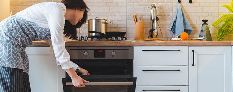 A woman reaches down to open the door of the under-bench oven in her modern white kitchen