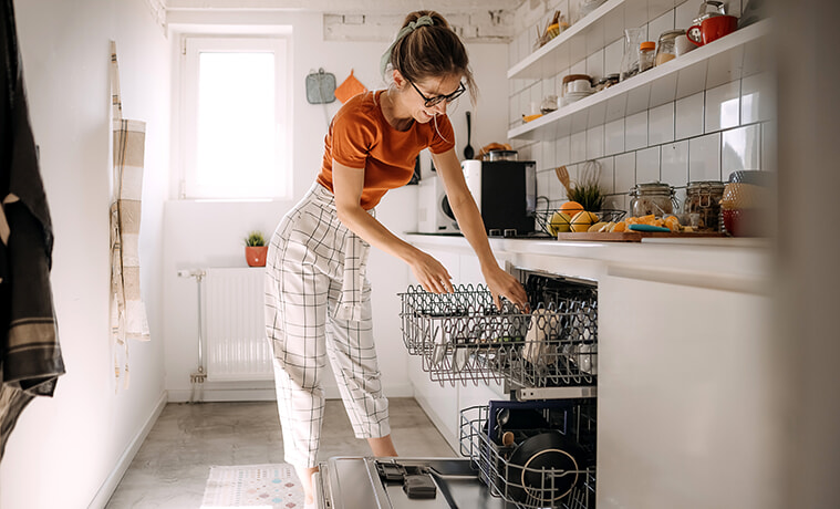 A young woman loads glasses into the top rack of a built-in dishwasher in her white galley-style kitchen.