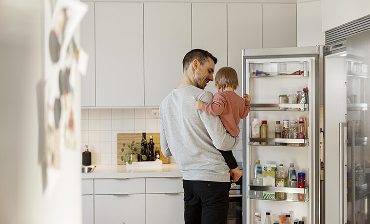 A father holds his toddler daughter while opening the door of his side-by-side fridge in a modern all-white kitchen.