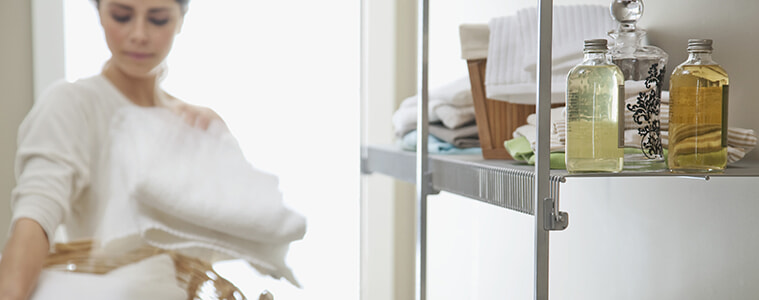 A woman takes a freshly folded white towel from the basket she is holding in her laundry with a neat and tidy shelving unit. 
