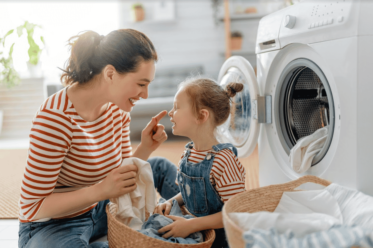 Mother and daughter laugh and smile while sitting on the floor in their laundry, with a basket of fresh clothes.