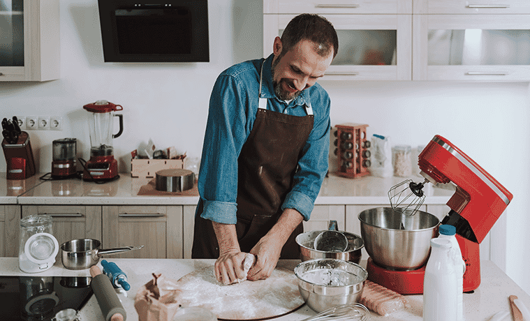 A man smiles and wears an apron while kneading a sweet pastry dough at his kitchen bench.