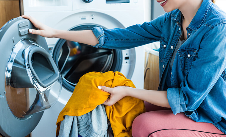 A smiling young woman removes freshly dried clothes from the heat pump dryer in her laundry.