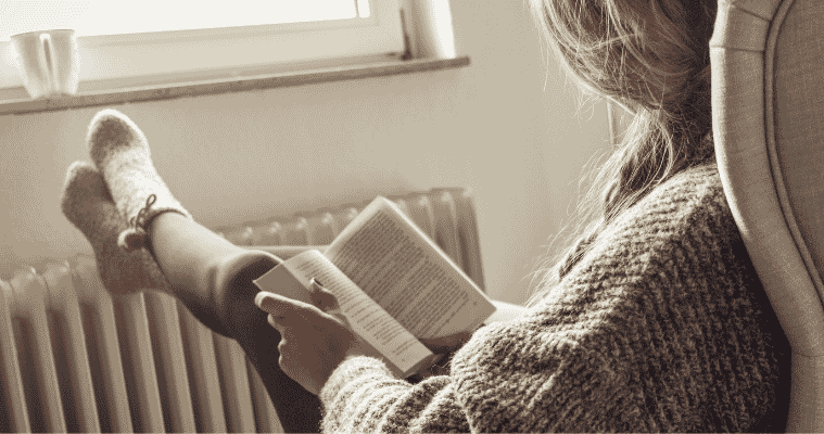 Woman reading a book with feet resting on a portable radiator.